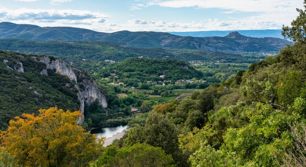 Magnifique vue en hauteur d'une vallée verdoyante du Vivarais d'Ardèche, avec des monts surplombants la photographie. Un cours d'eau, l'Ardèche, en contre bas, et un petit village au centre de toute cette nature.